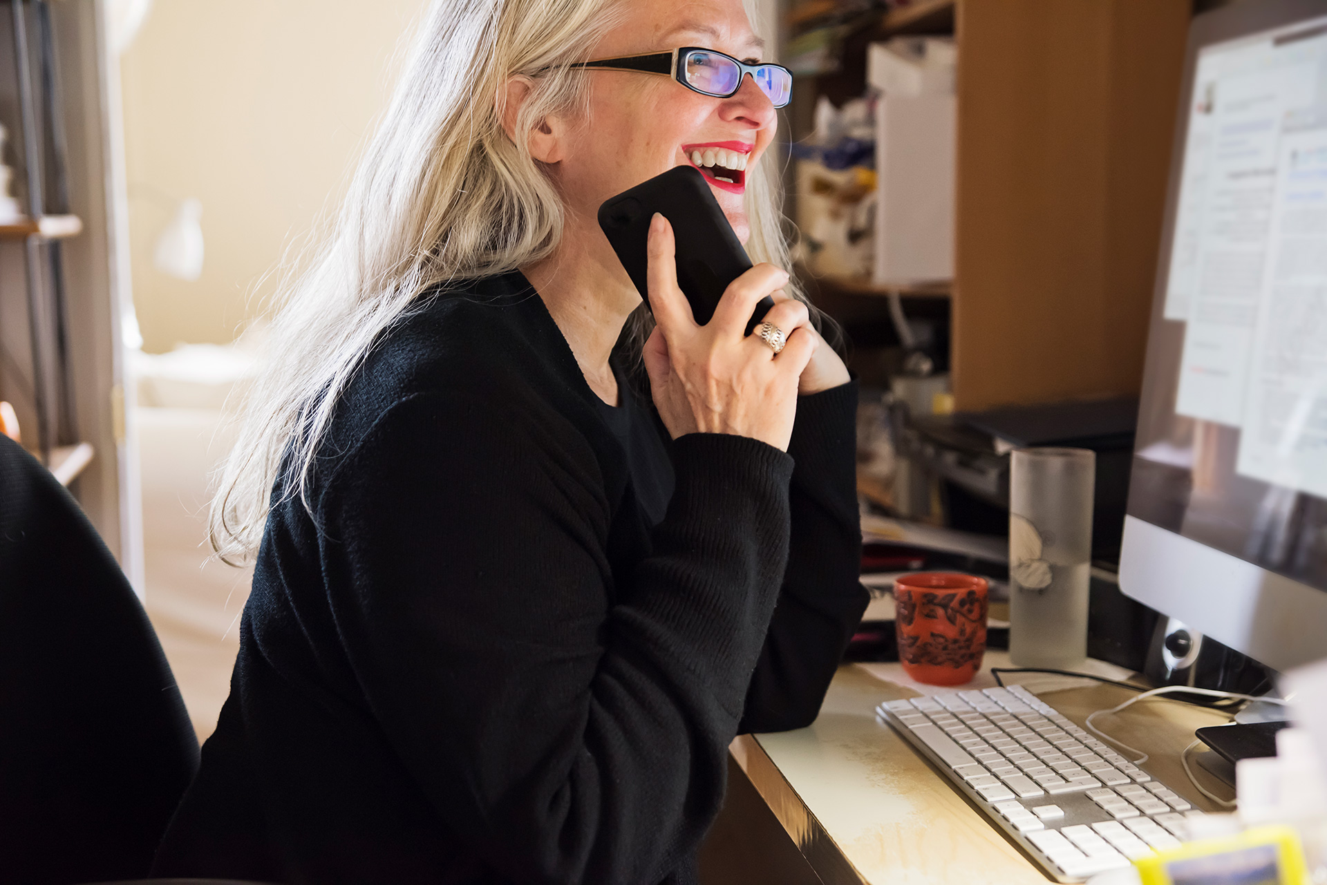 A woman sitting at a desk in front of a computer, laughing while she's on the phone.