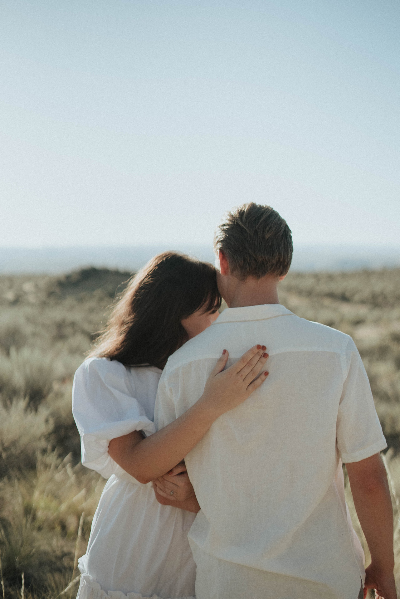 A couple wearing all white clothing embracing each other in a field of grass. 