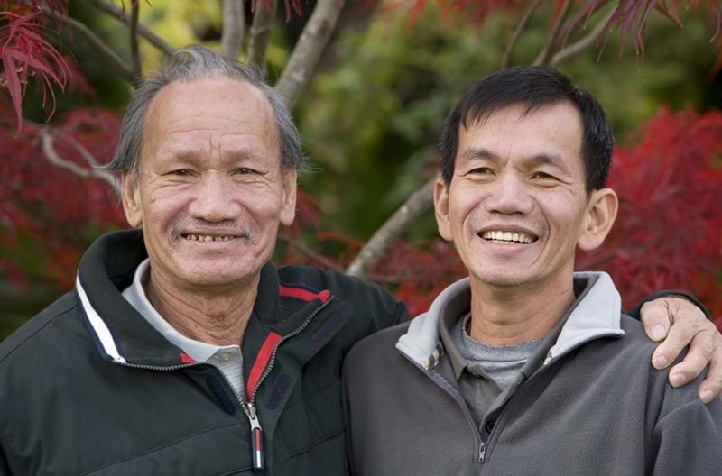An Asian adult standing with their parent, arms wrapped around each others shoulders.